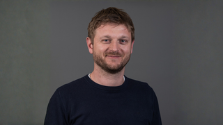 News Porträt eines lächelnden Mannes mit Bart vor grauem Hintergrund