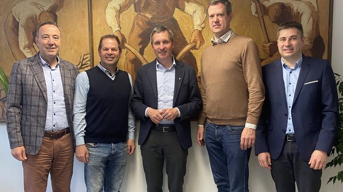 Primo incontro dei vertici di lvh.apa con l’assessore Luis Walcher Cinque uomini in abbigliamento formale davanti a dipinti murali con scene di lavoro