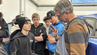 News Lehrer erklärt Schülern in Werkstatt etwas mit Tablet neben Auto