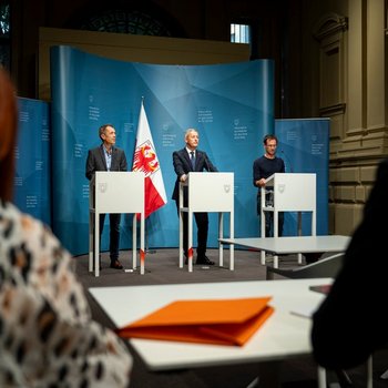 Kaminkehrer Drei Männer an Rednerpulten bei einer Pressekonferenz vor blauen Hintergründen und Flagge.