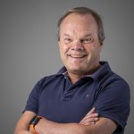 Das lvh-Team Lächelnder Mann mit blauem Poloshirt und orangener Smartwatch vor grauem Hintergrund