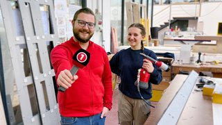 News Mann mit Mikrofon interviewt Handwerkerin mit Heißluftpistole in Werkstatt