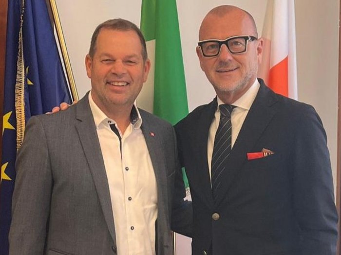 Kunst auf dem Rathausplatz – und ein Gespräch darüber, was sie auslöst Zwei Männer in Anzügen lächeln vor drei Flaggen in einem Innenraum