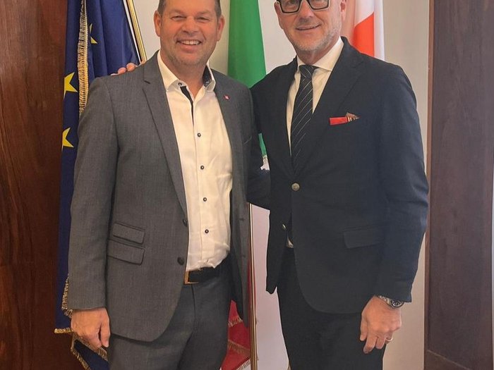 Arte in Piazza Municipio – e un dialogo sulle emozioni suscitate Due uomini in abiti sorridono davanti a tre bandiere in una stanza interna