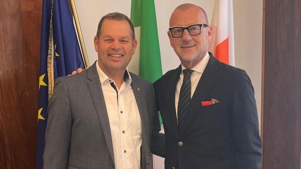 Zwei Männer in Anzügen lächeln vor drei Flaggen in einem Innenraum