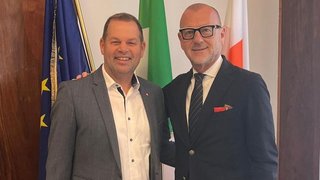 News Zwei Männer in Anzügen lächeln vor drei Flaggen in einem Innenraum