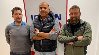 News Drei Männer stehen lächelnd vor einer weißen Wand mit rotem und blauem Banner
