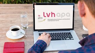 News Mann arbeitet am Laptop mit LVH APA Webseite, Kaffeetasse und Glas Wasser auf Tisch