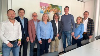 News Acht Personen stehen nebeneinander in einem modernen Besprechungsraum