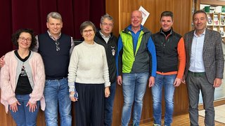 News Sieben Erwachsene stehen in einem Raum vor einer Holzwand mit rotem Vorhang