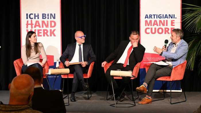"La città che desideri": un incontro per il futuro di Bolzano Discussione in panel con quattro partecipanti davanti a striscioni su artigianato e mestiere