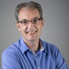 Maschinenschnitzer Porträt eines lächelnden Mannes mit Brille und blauem Hemd vor grauem Hintergrund