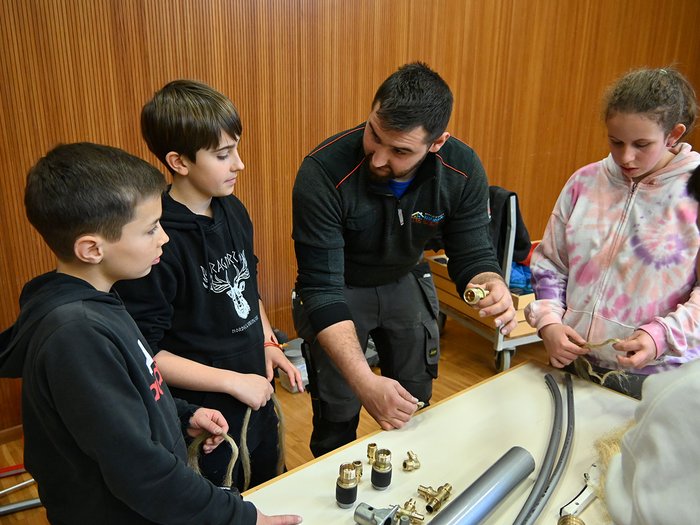 Handwerk zum Anfassen – 29. Tag des Handwerks in Jenesien Erwachsener zeigt Kindern Installation von Rohrleitungsteilen auf Tisch
