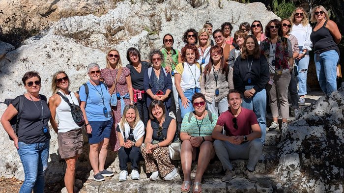 Sicilia – Sole, classe e donne forti: le donne di lvh.apa protagoniste di un viaggio pieno di stimoli Foto di gruppo di 27 persone davanti a una parete rocciosa con sole
