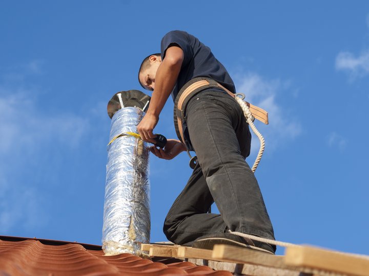 Kaminkehrer Handwerker mit Sicherheitsgurt repariert Schornstein auf einem Dach bei klarem Himmel