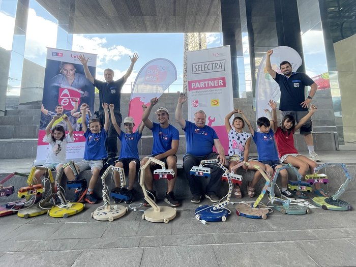 Projektwoche „Bunte Lichtermacher“: die Kinder mit den Handwerkern Herbert Kasal (links oben stehend), Alexander Morandell (rechts oben stehend), Daniel Rella und Herbert Bonora (in der Mitte sitzend). © lvh.apa Kinder und Erwachsene mit gebastelten elektrischen Lampen sitzen vor Bannern unter einem Dach
