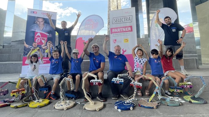 Projektwoche „Bunte Lichtermacher“: die Kinder mit den Handwerkern Herbert Kasal (links oben stehend), Alexander Morandell (rechts oben stehend), Daniel Rella und Herbert Bonora (in der Mitte sitzend). © lvh.apa Kinder und Erwachsene mit gebastelten elektrischen Lampen sitzen vor Bannern unter einem Dach