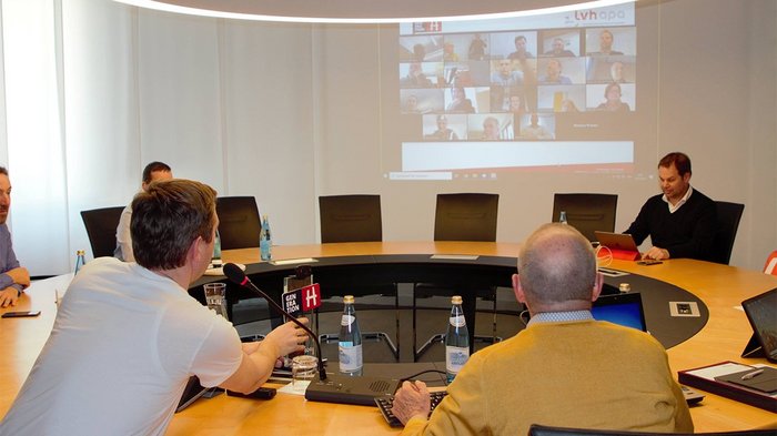 „COVID-19 hat uns in die Knie gezwungen, wir werden aber wieder aufstehen!“ Menschen in einem Konferenzraum bei einer hybriden Videokonferenz mit Bildschirmprojektion