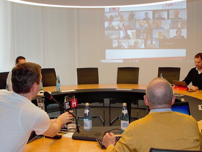 „COVID-19 hat uns in die Knie gezwungen, wir werden aber wieder aufstehen!“ Menschen in einem Konferenzraum bei einer hybriden Videokonferenz mit Bildschirmprojektion