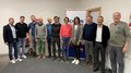 Frauenduo führt Handwerkergruppe an Elf Personen posieren in einem Büro vor einem Schild mit Schriftzug Handwerk