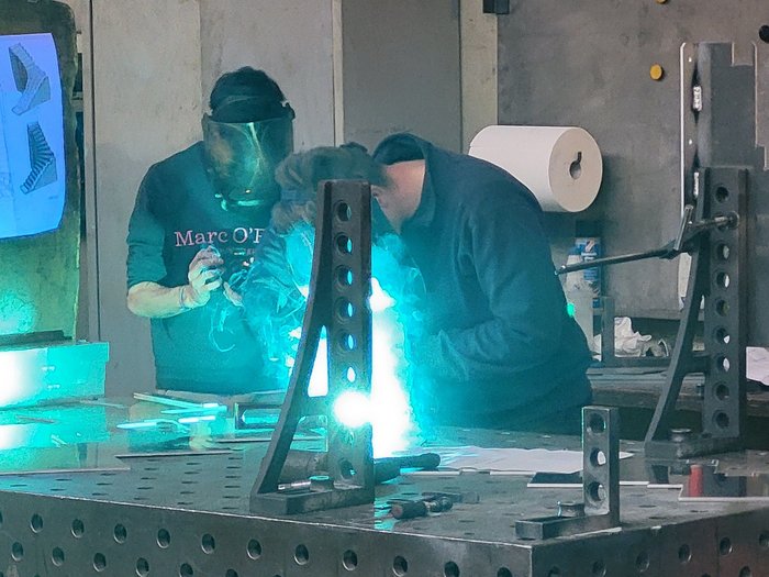 „Was du baust, bewegt die Welt“ Zwei Personen schweißen mit Schutzmaske an Metallteile in Werkstatt