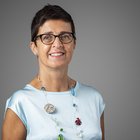 Elisabeth Waldthaler Ritratto di una donna sorridente con capelli corti, occhiali e maglia azzurra su sfondo grigio