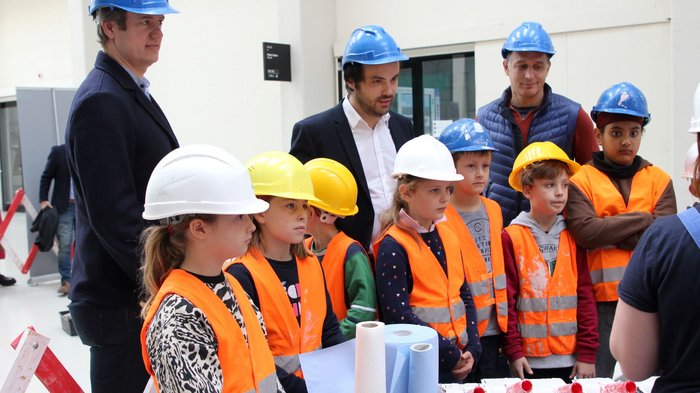Vivere una giornata in cantiere Bambini con caschi protettivi e giubbotti arancioni a una dimostrazione edilizia con adulti