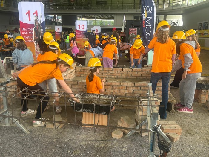 Nachwuchsinitiative „Abenteuer Handwerk“ der Intercable Arena Kinder in orangenen Shirts und gelben Helmen mauern Ziegel im Workshop