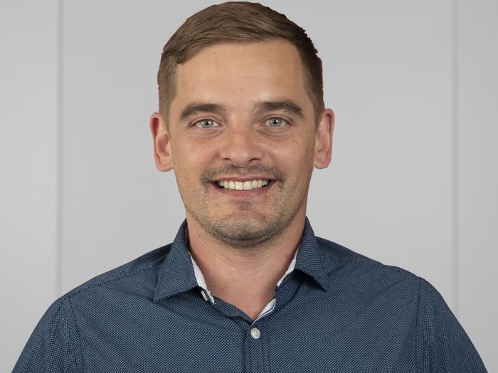 Kleine Betriebe als große Überlebenskünstler Porträt eines lächelnden Mannes in blauem Hemd vor neutralem Hintergrund