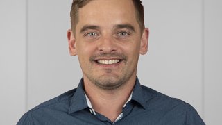 News Porträt eines lächelnden Mannes in blauem Hemd vor neutralem Hintergrund