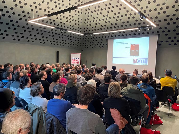 „Multitalent Beton“: ein voller Erfolg! Viele Menschen verfolgen eine Präsentation in einem modernen Raum mit perforierter Decke.