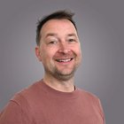 Kfz-Mechatroniker Lächelnder Mann mit braunem Pullover vor grauem Hintergrund