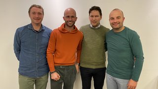 News Vier Männer stehen eng nebeneinander vor einer weißen Wand und lächeln