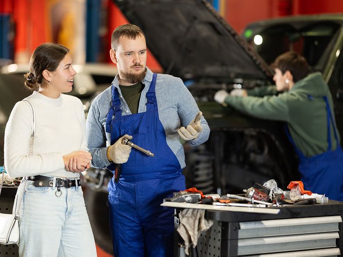 Prämie für Lehrbetriebe: Unterstützung für Ausbildung im Handwerk Mechaniker erklärt Kundin Autoreparatur in Werkstatt mit geöffnetem Motorhaube