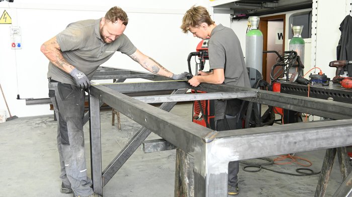 Ab sofort fahren auch Lehrlinge günstiger Zwei Männer arbeiten an einem Metallrahmen in einer Werkstatt