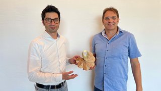 © Studiooberhauser Zwei Männer präsentieren ein hölzernes fächerförmiges Produkt vor weißer Wand