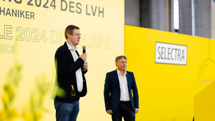 JV Elektrotechniker Zwei Männer sprechen auf einer Veranstaltung vor gelbem Hintergrund mit Selectra-Logo