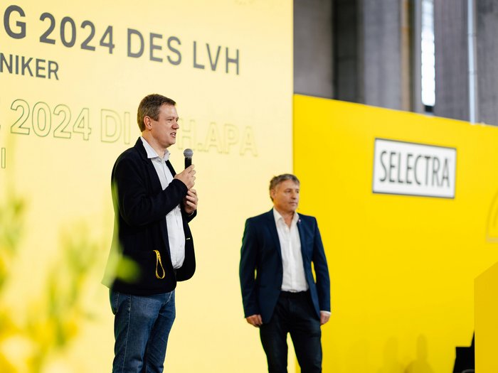 JV Elektrotechniker Zwei Männer sprechen auf einer Veranstaltung vor gelbem Hintergrund mit Selectra-Logo