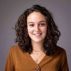 Lisa Huber Giovane donna sorridente con capelli ricci e maglia marrone su sfondo grigio