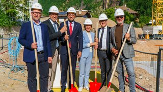 News Sechs Personen mit Helmen und Schaufeln auf einer Baustelle vor Bergen