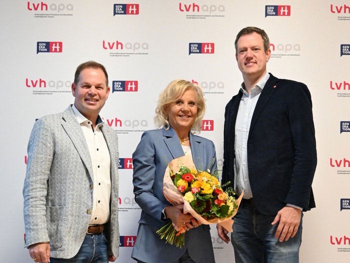 Angelika Wiedmer Perkmann rückt als erste Frau in die lvh-Verbandsspitze auf Drei Personen posieren vor einem Hintergrund mit Logos, eine hält einen Blumenstrauß